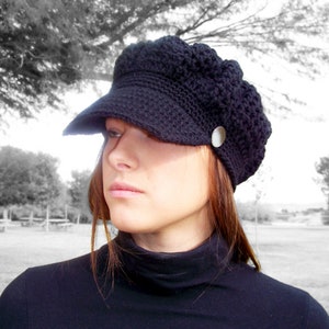 May include: A black crocheted newsboy cap with a visor and a silver button. The cap is worn by a person with long brown hair.