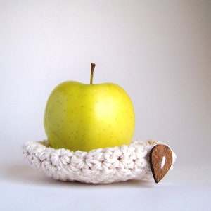 A yellow apple resting on a white hand crocheted apple cozy with a brown leaf-shaped button attached to its side.