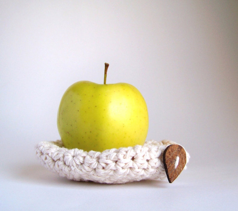 A yellow apple resting on a white hand crocheted apple cozy with a brown leaf-shaped button attached to its side.