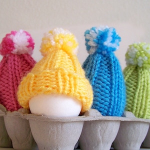 May include: Four eggs are decorated with knitted hats in a variety of colours, including pink, yellow, blue, and green. The hats have white pom-poms on top. The eggs are sitting in a cardboard egg carton.