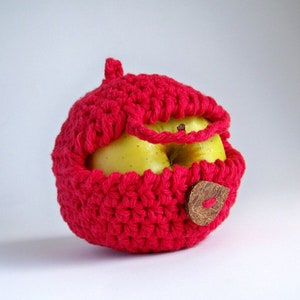 Puede incluir: Una funda de manzana de punto de ganchillo rojo con un botón de madera. La funda cubre una manzana amarilla.