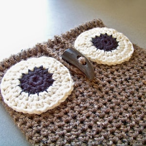 Puede incluir: Búho de crochet marrón con ojos blancos de crochet y una nariz de botón negro. El búho tiene un botón marrón en forma de cuerno en la parte superior.
