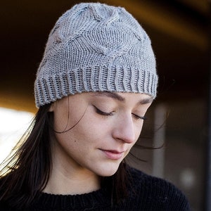 Young woman wearing a light gray knitted Seagulls Hat with a cable knit pattern, eyes closed and head tilted down, featuring dark brown hair peeking out, set against a softly blurred background. This listing is for an easy hat knitting pattern.