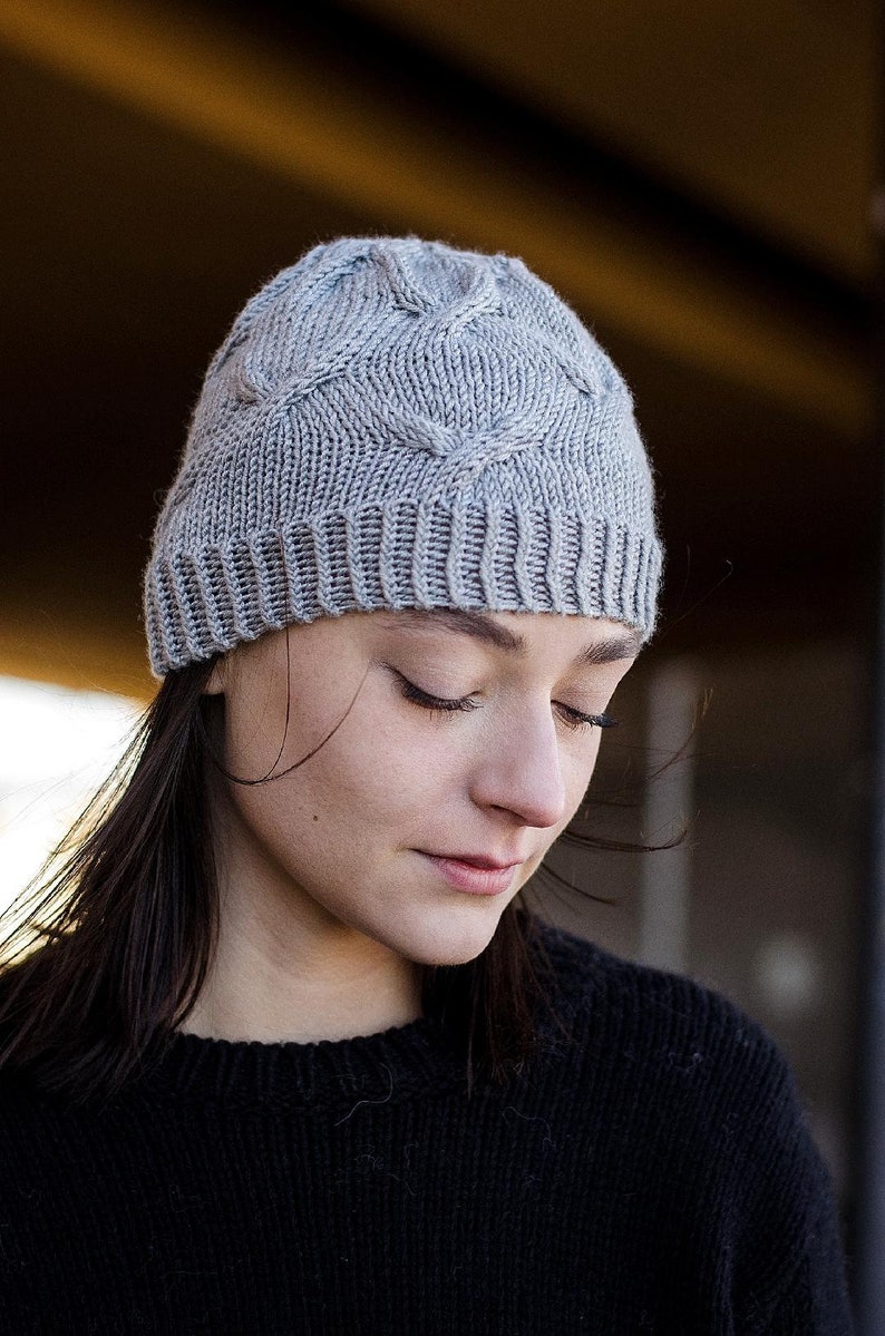 Young woman wearing a light gray knitted Seagulls Hat with a cable knit pattern, eyes closed and head tilted down, featuring dark brown hair peeking out, set against a softly blurred background. This listing is for an easy hat knitting pattern.