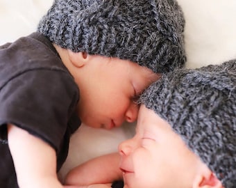 May include: Two babies wearing gray knit hats are nestled together, their faces close. The hats have a textured cable knit design. The babies are wearing dark gray shirts. The image is a close-up, capturing the peaceful scene.