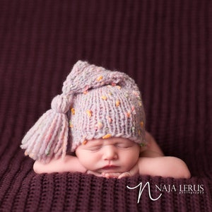 May include: A newborn baby wearing a pink knitted hat with a long tassel. The hat has a multi-coloured fleck pattern. The baby is sleeping with their arms crossed in front of them.