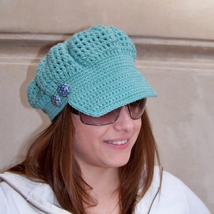 May include: A teal blue crocheted hat with a visor and two decorative buttons. The hat is worn by a person with long brown hair.