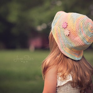 A young girl with long wavy hair wearing a pastel-colored cotton crochet sun hat with a pink flower embellishment. The background is a blurred green grassy area with trees.