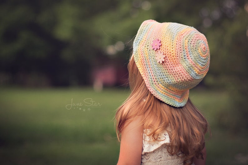 A young girl with long wavy hair wearing a pastel-colored cotton crochet sun hat with a pink flower embellishment. The background is a blurred green grassy area with trees.