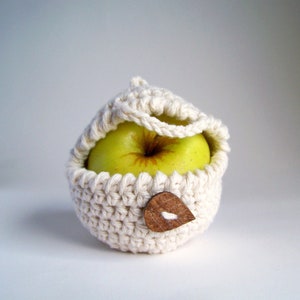 An off-white crochet apple cozy with a brown leaf-shaped wooden button fastened around a green apple on a white background.