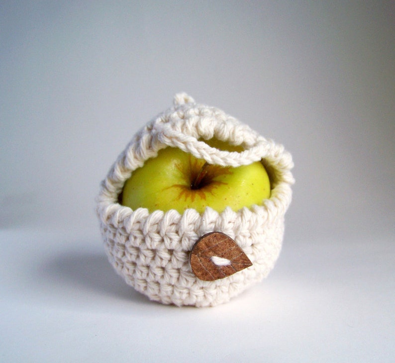 An off-white crochet apple cozy with a brown leaf-shaped wooden button fastened around a green apple on a white background.