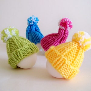 May include: Four white eggs are topped with colourful knitted hats. The hats are green, pink, blue, and yellow with white pom-poms.