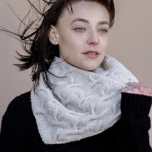 May include: A woman wearing a white knitted cowl neck scarf with a cable knit pattern. The scarf is worn over a black sweater.