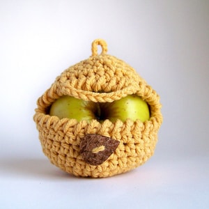 Puede incluir: Una funda de manzana de ganchillo amarilla con un detalle de hoja marrón. La funda está diseñada para adaptarse a una sola manzana.