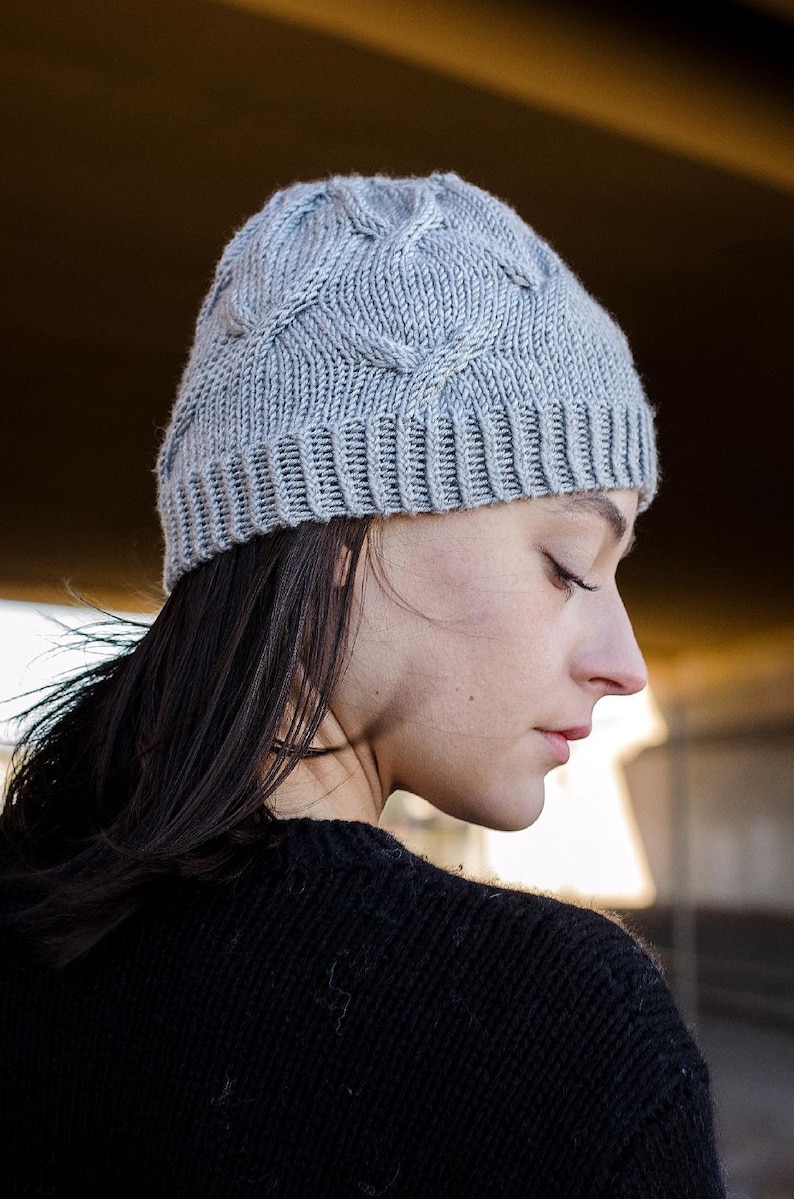 Side profile of a young woman wearing a light gray knit Seagulls Hat and a black sweater, standing under a bridge. This is an easy beanie knitting pattern.