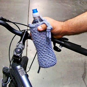 May include: A gray crocheted water bottle holder attached to a bicycle handlebar. The bottle holder is made of a soft, knitted fabric and has a blue plastic water bottle inside.