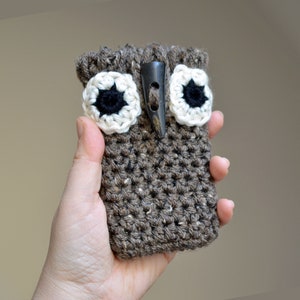 May include: A brown crocheted owl-shaped phone case with white and black eyes and a brown button.
