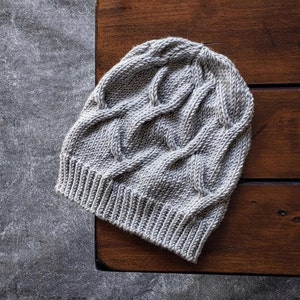 A gray knit Seagulls Hat with a ribbed brim and cable stitch laid flat on a dark brown wooden table. it is an easy knitting pattern.