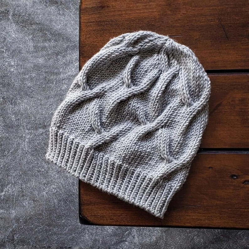 A gray knit Seagulls Hat with a ribbed brim and cable stitch laid flat on a dark brown wooden table. it is an easy knitting pattern.