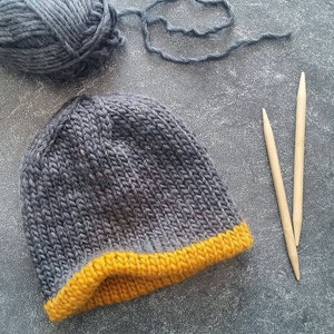 Könnte beinhalten: Eine graue Strickmütze mit gelbem Rand. Die Mütze liegt auf einer grauen Oberfläche mit zwei Holzstricknadeln.
