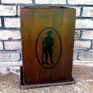 May include: A wooden box with a dark brown finish. A black silhouette of a person in colonial attire is inside an oval frame. The box has a rectangular base and is set against a brick wall.