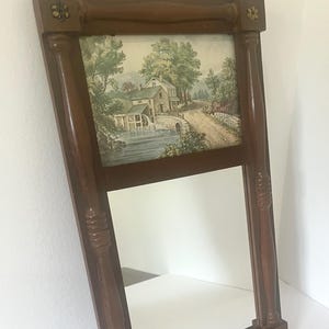May include: A vintage wooden mirror with a landscape print at the top. The mirror has a dark brown frame with decorative floral accents. The landscape print depicts a rural scene with a house, water, and trees.