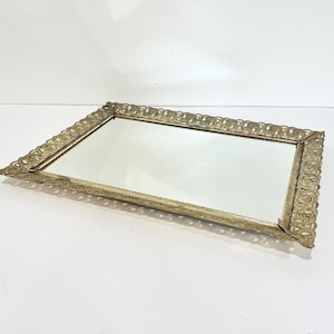 May include: A rectangular, gold-toned vanity tray with a mirrored surface. The tray features an ornate, openwork filigree border with a scalloped edge. The tray is set against a white background, showcasing its decorative design.