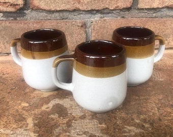 May include: Three white ceramic mugs with brown and tan bands around the top. The mugs have rounded handles.