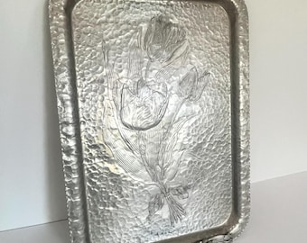 May include: A rectangular, silver-colored metal tray with a hammered texture. The tray features an embossed design of tulips and leaves. The edges are adorned with floral and scroll details, and the handles have a decorative design.