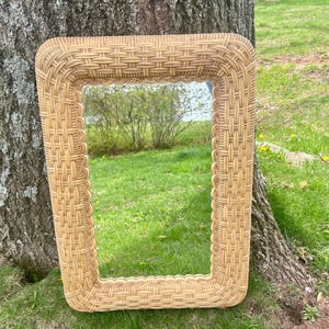 May include: A rectangular wicker mirror with a woven frame. The mirror is leaning against a tree trunk in a grassy area.