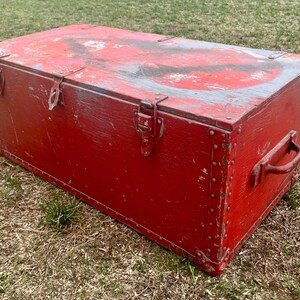 May include: A large, red wooden storage chest with metal hardware and a worn, distressed finish. The chest has a hinged lid with two metal clasps and a metal handle on the side.