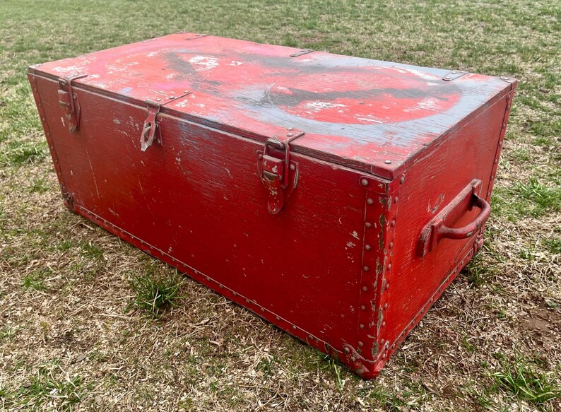 May include: A large, red wooden storage chest with metal hardware and a worn, distressed finish. The chest has a hinged lid with two metal clasps and a metal handle on the side.