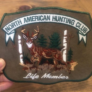 May include: A brown suede patch with green embroidered trim. The patch features a white banner with the text "North American Hunting Club" and a smaller white banner with the text "Life Member". A brown and white deer is embroidered in the center of the patch.