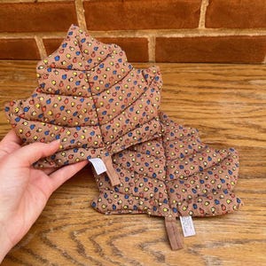May include: Brown leaf-shaped quilted potholder with a floral pattern in blue, yellow, and red. The potholder has a tag attached. The background is a wooden surface and a brick wall.