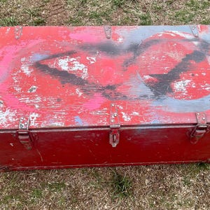 May include: A red wooden storage chest with a hinged lid and metal clasps. The chest has a distressed paint finish with faded blue and black paint accents.