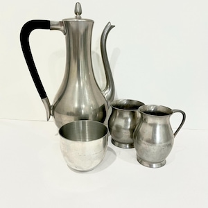 May include: A silver-toned metal coffee service set consisting of a tall, slender coffee pot with a black handle and lid, a small creamer with a handle, and two matching cups. The pieces have a smooth, metallic finish and a simple, functional design.