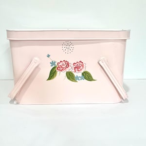 May include: A rectangular light pink metal basket with a hinged handle on each side. The front features a painted floral design of two pink roses with green leaves and small blue flowers, positioned below a small circular pattern of ventilation holes.