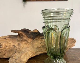 May include: A green glass vase with a unique, fluted design. The vase has a wide base and a narrow neck. It is sitting on a wooden surface.