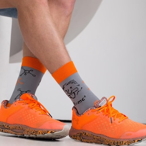 Könnte beinhalten: Ein Paar orangefarbene und graue Sportsocken mit einer Comicfigur und der Gleichung "E=mc^2" auf einem Socken und einer chemischen Struktur auf dem anderen Socken. Die Socken werden mit orangefarbenen und schwarzen Sportschuhen getragen.