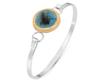 May include: A silver bangle bracelet featuring a round blue and gold ceramic charm. The charm has a gold rim and a blue centre with a darker blue swirl pattern.