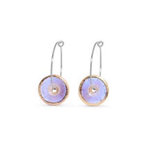 May include: A pair of ceramic hoop earrings. Each earring features a light purple disc with a gold-colored rim and a silver-colored hoop. The earrings are set against a white background.