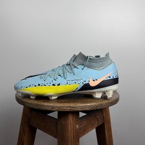May include: Light blue football boots with a black and yellow patterned design. The boots have a white sole and a pink Nike swoosh logo. The text "PHANTOM" is printed on the side of the boots.