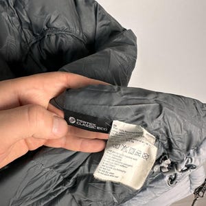 Puede incluir: Primer plano de una chaqueta acolchada gris con capucha. La chaqueta tiene una etiqueta Pertex Classic Eco y una etiqueta con instrucciones de cuidado. La chaqueta est&aacute; hecha de poli&eacute;ster reciclado y plum&oacute;n de pato.