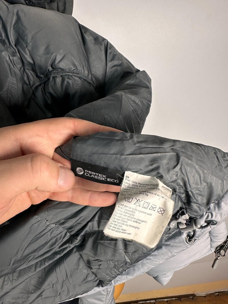 Puede incluir: Primer plano de una chaqueta acolchada gris con capucha. La chaqueta tiene una etiqueta Pertex Classic Eco y una etiqueta con instrucciones de cuidado. La chaqueta est&aacute; hecha de poli&eacute;ster reciclado y plum&oacute;n de pato.