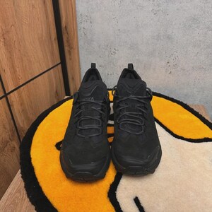 May include: A pair of black athletic shoes with textured uppers and black laces. The shoes are displayed on a yellow and white rug with a black border, set against a wooden wall.