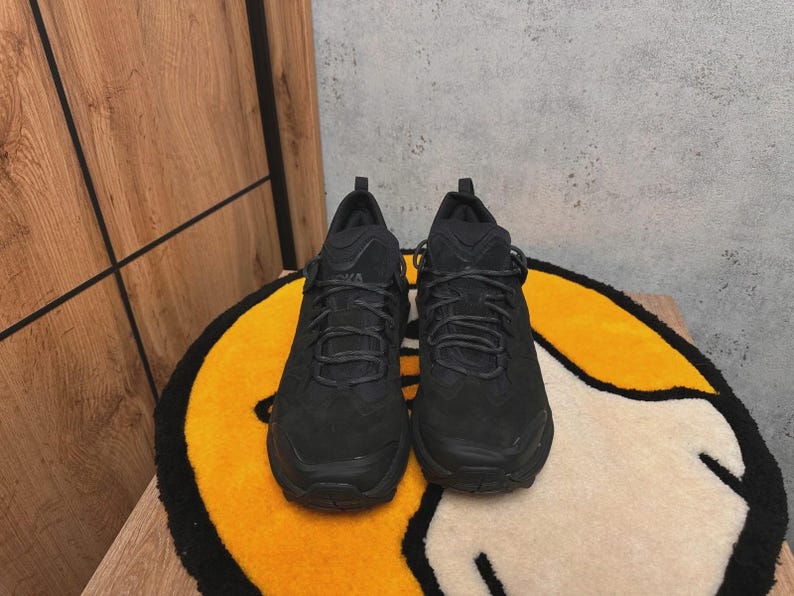 May include: A pair of black athletic shoes with textured uppers and black laces. The shoes are displayed on a yellow and white rug with a black border, set against a wooden wall.