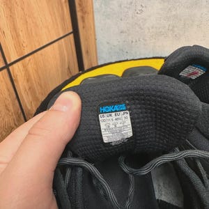 May include: Close-up of a black athletic shoe with gray laces. The shoe has a label with the brand name "HOKA ONE" and size information: US 12D (11.5 46%) 30. The shoe is on a yellow surface.