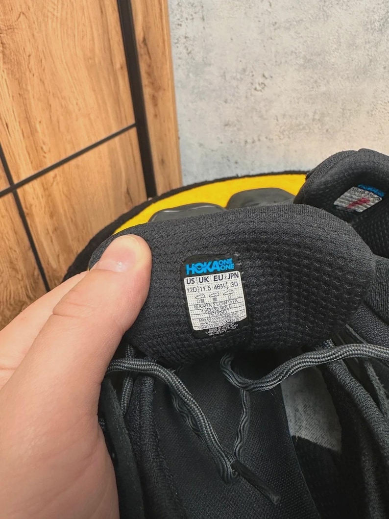 May include: Close-up of a black athletic shoe with gray laces. The shoe has a label with the brand name "HOKA ONE" and size information: US 12D (11.5 46%) 30. The shoe is on a yellow surface.