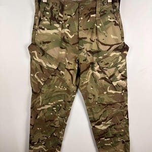 Könnte beinhalten: Tarnfarbene Cargohose in Grün-, Braun- und Beigetönen. Die Hose hat mehrere Taschen, einen Knopfverschluss und einen Kordelzug in der Taille. Die Hose hängt an einem Kleiderbügel.