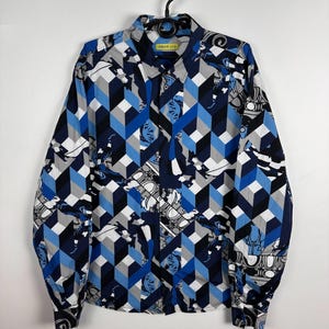 May include: A long-sleeved button-down shirt with a geometric cube pattern in shades of blue, gray, and black. The shirt also features a repeating design of white and gray decorative elements. The label reads "VERSACE JEANS".